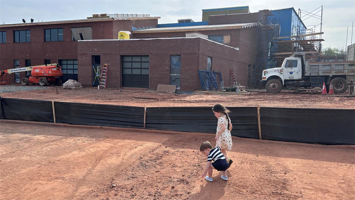Active construction site of a large brick building in the background of picture surrounded by a black plastic fence, kids in foreground of picture playing in dirt